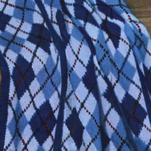 Può includere: Una coperta lavorata a maglia con un classico motivo a rombi nei toni del blu navy, azzurro e bianco. Il design è composto da forme a diamante intrecciate, accentuate da sottili linee diagonali rosso scuro che attraversano il motivo blu e bianco.
