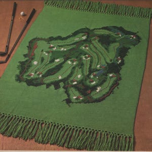 May include: Green knitted rug with a detailed design of a golf course. The rug has fringe around the edges.