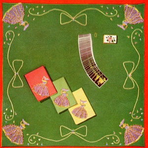 May include: A green felt table cloth with a red border. The table cloth is decorated with embroidered flowers and bows. There are four embroidered napkins with purple and gold designs. A deck of cards is fanned out on the table. There is a small box with a rose design on the table.