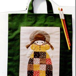 Könnte beinhalten: Grüne Tragetasche mit Stoffpuppen-Applikation. Die Puppe hat einen braunen Hut, Zöpfe und ein gelbes Kleid mit einem Patchwork-Rock. Zwei Buntstifte liegen neben der Tasche.