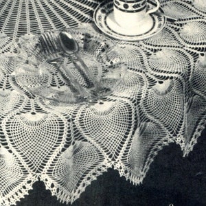 May include: A white crocheted tablecloth with a delicate, lacy pattern featuring a repeating design of fan-shaped motifs. The tablecloth is laid out on a dark surface and has a scalloped edge.