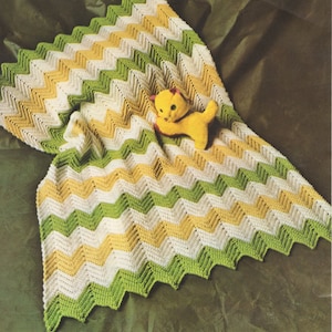 May include: A crocheted baby blanket with a chevron pattern in green, white and yellow. A yellow stuffed animal is lying on the blanket.