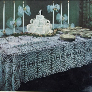 May include: A white crocheted tablecloth with a geometric pattern, set for a wedding reception. The table is decorated with a white wedding cake, candles, and white bows.
