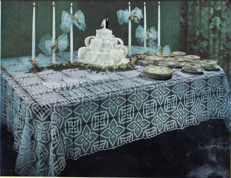 May include: A white crocheted tablecloth with a geometric pattern, set for a wedding reception. The table is decorated with a white wedding cake, candles, and white bows.