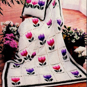 May include: A crocheted blanket and cushion set with a repeating tulip design in pink and purple on a white backdrop. The blanket has a black border. The set is displayed on a wooden chair.