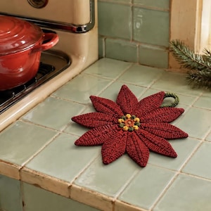 May include: A dark red, poinsettia-shaped crochet potholder featuring textured petals, a central cluster of small yellow and green knots, and a small green loop for hanging, designed to protect surfaces from hot cookware.