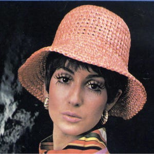 May include: A vintage image of a woman wearing a peach-colored woven sun hat with a wide brim. The hat has a perforated design. She is wearing gold hoop earrings and a colorful striped top. The background is dark.