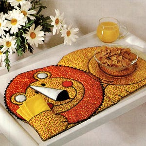 May include: A breakfast tray with a lion-shaped placemat, a glass bowl of cereal, and a glass of orange juice. The placemat is yellow with red floral accents and a lion face design. A bouquet of white daisies is in the background.