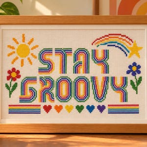 May include: A framed cross-stitch embroidery featuring the text STAY GROOVY in rainbow-coloured letters. The design includes a yellow sun, a rainbow shooting star, a red flower, a blue flower, and a row of colourful hearts and stripes.