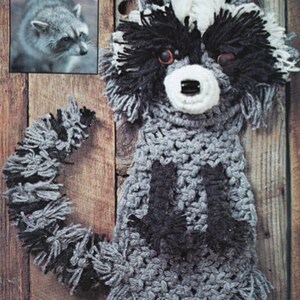 May include: A crocheted raccoon wall hanging with a gray and black color scheme. The raccoon has a white face with black eyes and a black nose.