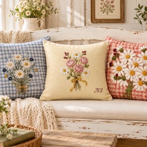 May include: Three square throw pillows featuring embroidered floral bouquets. The left pillow has a blue and white gingham pattern, the center is solid cream with the initials NS, and the right has a red and white gingham pattern.