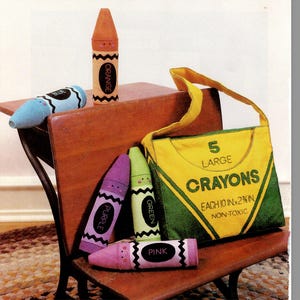 Puede incluir: Una colección de objetos con forma de crayones de colores, incluyendo un crayón naranja, azul, morado, verde y rosa. Una bolsa tote amarilla y verde dice "5 GRANDES CRAYONES CADA UNO 25.4 CM 6.35 CM NO TÓXICO".