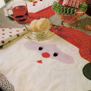 May include: A white table runner with a Santa Claus face design. The runner has a red border and a green and white polka dot pattern. There are white coconut candies in a glass bowl and a glass bowl filled with colorful candy.