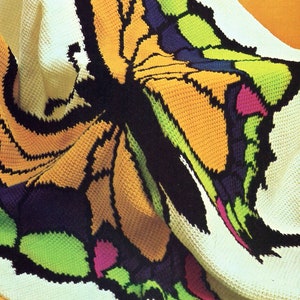 May include: A crocheted blanket featuring a colourful butterfly design. The butterfly is made with different coloured yarn, including orange, green, blue, and black. The blanket is white with a crocheted texture.