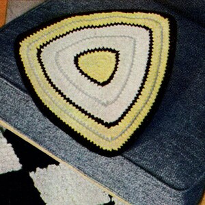 May include: A handmade, triangular crochet doily with a yellow, white, and black color scheme. The doily is placed on a blue upholstered chair. The doily's design features concentric triangles, with a black border and a yellow center.