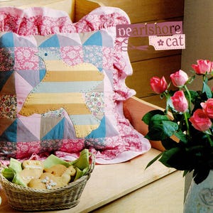 May include: A decorative pillow with a patchwork bird design in pastel colors, resting on a pink floral patterned pillow. The image includes a basket of cookies and a vase of pink roses. The text "pearlshore cat" is visible.
