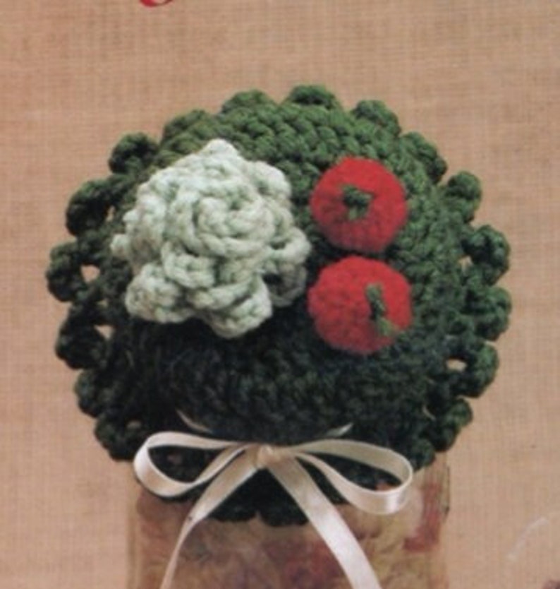 May include: A green crocheted jar lid cover with a white crocheted flower and two red crocheted tomatoes on top. A white satin ribbon is tied around the base of the lid.