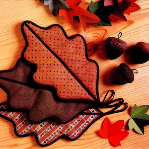 Può includere: Una collezione di decorazioni in tessuto a tema autunnale. L'immagine presenta tre tovagliette a forma di foglia nei toni del marrone e dell'arancione, con bordi neri. Sono inclusi anche tre ornamenti a forma di ghianda e diverse foglie di carta rosse e verdi.