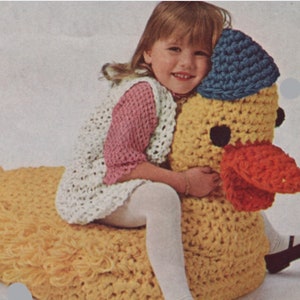 May include: A crocheted yellow duck toy with a red beak and blue hat. A young girl is sitting on the duck toy.