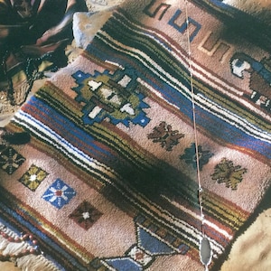 May include: A colorful woven rug with geometric patterns in shades of blue, green, brown, and white. The rug has a fringe border and is laid out on a sandy surface.