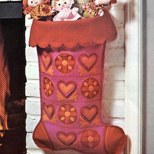 Peut inclure: Une chaussette de Noël en feutre rose avec un bord festonné et un motif de cœurs et de fleurs orange et jaunes. La chaussette est accrochée à un mur de briques blanches.