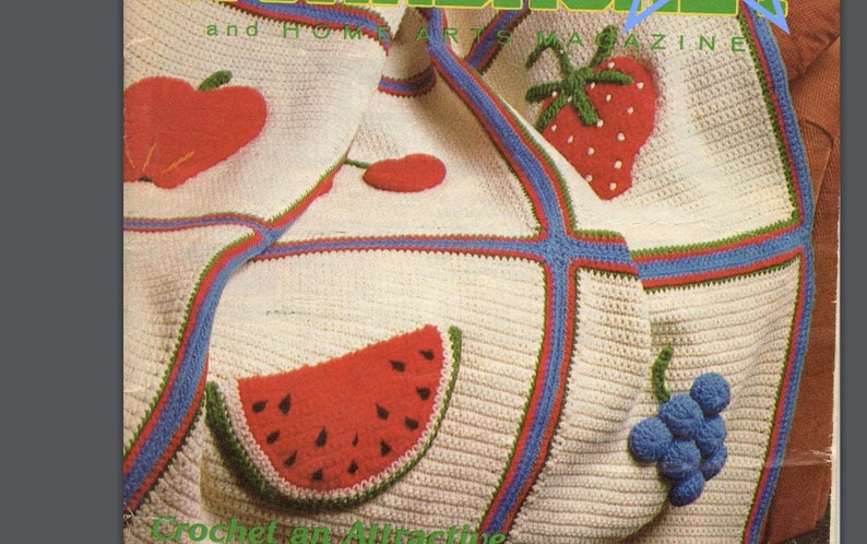 May include: A cream-colored crochet blanket featuring colorful fruit appliques including a red apple, a red strawberry with white seeds, a red watermelon slice with black seeds, and a cluster of blue grapes, all bordered by blue and green yarn.