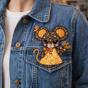 Puede incluir: Chaqueta vaquera azul claro con un parche decorativo de un ratón de dibujos animados. El ratón tiene un cuerpo con estampado floral, ojos grandes y sostiene trigo. El parche está en el bolsillo delantero.