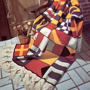 Op de afbeelding: Een gehaakte deken met een nautisch thema met verschillende kleurrijke seinvlaggen. De deken heeft een franje rand en is over een houten stoel gelegd.