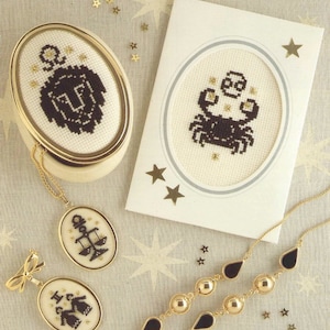 May include: A set of zodiac-themed jewellery and a card with a gold and black crab design. The jewellery includes a necklace with a gold pendant featuring a black Libra scale design, a pair of earrings with a gold bow and a black Gemini design, and a gold bracelet with black teardrop-shaped beads.