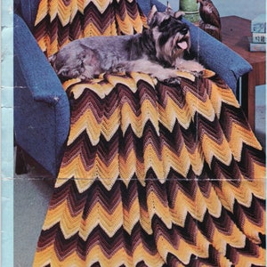 May include: A handmade, chevron-patterned blanket in shades of brown, yellow, and cream is draped over a blue armchair. A dog is lying on the blanket. The blanket's design creates a zig-zag pattern. The blanket is a cozy home decor item.