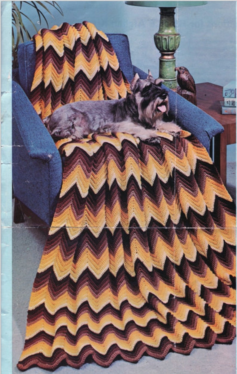 May include: A handmade, chevron-patterned blanket in shades of brown, yellow, and cream is draped over a blue armchair. A dog is lying on the blanket. The blanket's design creates a zig-zag pattern. The blanket is a cozy home decor item.
