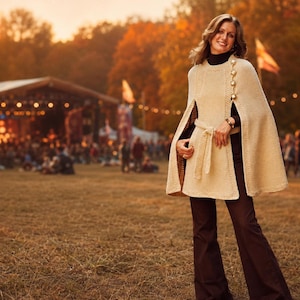 May include: A cream-colored knit cape featuring a round neckline, a row of gold-toned buttons along the right shoulder, and a matching tie belt at the waist. The garment is worn over a black mock-neck top and dark brown flared trousers.