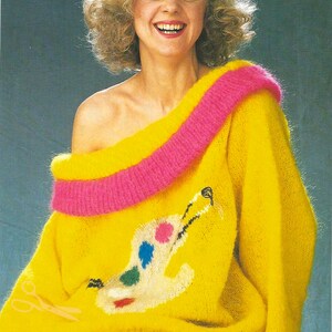 May include: A yellow fuzzy sweater with a pink trim around the neckline. The sweater has a colorful paint palette design on the front.