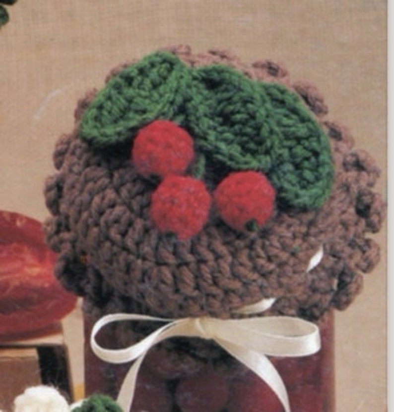 May include: A brown crocheted jar lid with a white ribbon and a crocheted cherry design on top. The cherries are red and the leaves are green.