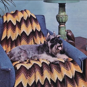 May include: A gray and black dog rests on a blue armchair, draped with a handmade chevron-patterned blanket. The blanket features a zig-zag design in shades of yellow, brown, and dark brown. A green lamp and a wooden bird statue are visible in the background.