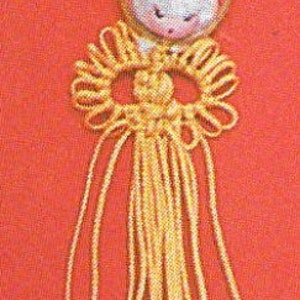 May include: A yellow macrame knot with a white angel charm hanging from a gold ring. The angel has a white face with black eyes and a pink mouth.