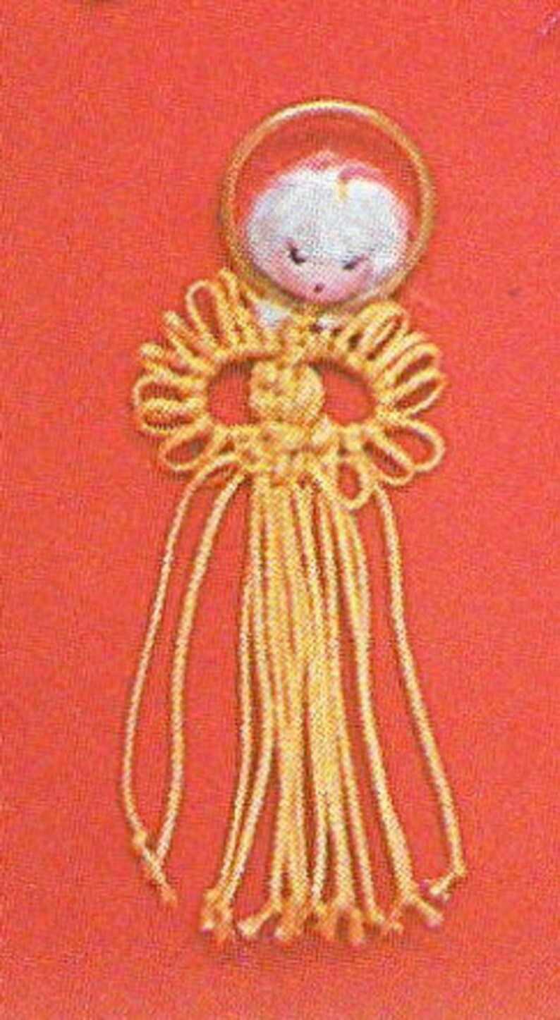 May include: A yellow macrame knot with a white angel charm hanging from a gold ring. The angel has a white face with black eyes and a pink mouth.