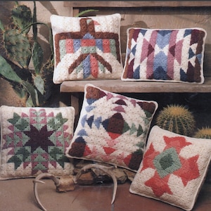 Puede incluir: Cinco fundas de cojín de ganchillo con patrones geométricos en tonos de marrón, rosa, verde, azul y blanco. Los cojines están dispuestos en un estante de madera con una planta de cactus en el fondo.