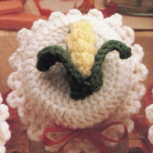 May include: White crocheted ornament with a yellow and green corn design. The ornament is tied with a red ribbon.