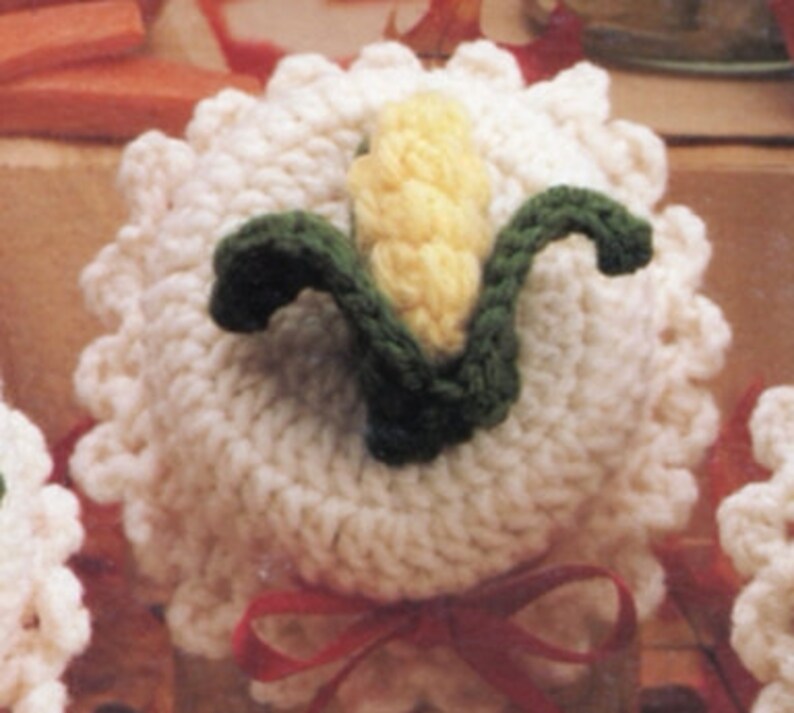 May include: White crocheted ornament with a yellow and green corn design. The ornament is tied with a red ribbon.