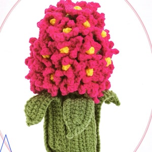 May include: A crocheted hyacinth flower with pink petals and yellow centers. The flower is in a green crocheted pot with leaves.
