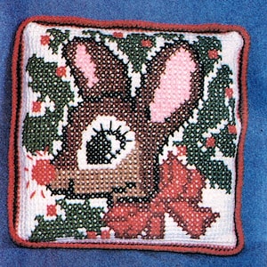 May include: A square, cross-stitched pillow featuring a reindeer design. The reindeer has brown fur, pink ears, and a red bow. Green holly and red berries surround the reindeer on a white background, with a red border. A festive, holiday-themed item.