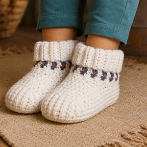 May include: Off-white crocheted slipper boots with a ribbed cuff and a decorative band with a floral pattern in green and pink. The boots are on a beige rug.