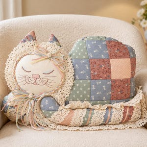 May include: A cat-shaped decorative pillow featuring a patchwork body in muted shades of blue, green, and pink. The face is cream-colored with embroidered closed eyes and whiskers, framed by cream lace trim and accented with pastel ribbon bows.
