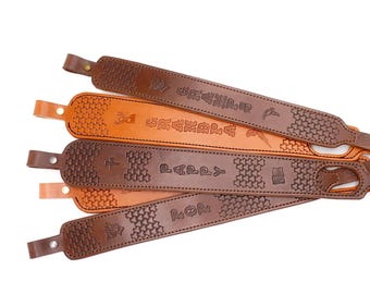 May include: A collection of leather rifle slings in various shades of brown and tan. Each sling features embossed designs and text, including a cross, deer, and other symbols. The slings have a textured pattern and a loop for attachment.