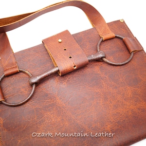 May include: A brown leather crossbody bag with a metal bit buckle closure. The bag has a long strap and a shorter handle. The bag is made by Ozark Mountain Leather.