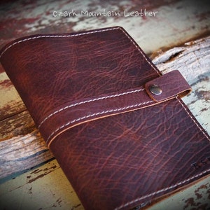 May include: A brown leather journal with a strap closure and a white stitch detail. The journal has a textured surface and the initials "R.G." are embossed on the front cover.