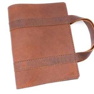 May include: A brown leather book cover with a handle, featuring two horizontal straps and stitched detailing. The initials "LBK" are visible on the lower right corner. The cover is designed to hold a book.