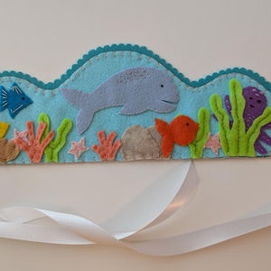 May include: A felt crown with an ocean theme. The crown is light blue with a scalloped edge and features felt cutouts of a whale, fish, octopus, coral, and seaweed. White ribbon is attached to the sides.