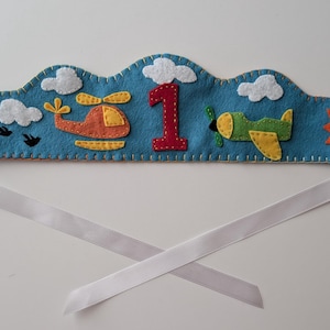 May include: A blue felt crown with a cloud-like shape, featuring appliqued designs of a hot air balloon, helicopter, airplane, sun, clouds, and the number 1. White ribbon ties are attached for securing the crown.